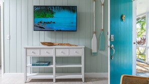 Ein Zimmer mit einer türkisblauen Tür, einem weißen Konsolentisch mit Schubladen, einem an der Wand montierten Fernseher, der eine Strandansicht zeigt, und einem an der Wand lehnten Paddel.