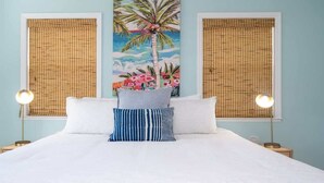 Een bed met witte beddengoed, twee kussens, een blauw-wit gestreept kussen, en een schilderij van een palmboom en bloemen aan de muur.
