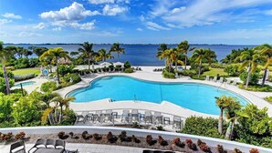 Une grande piscine incurvée, entourée de palmiers et de chaises longues, offrant une vue sur la mer et un ciel dégagé.