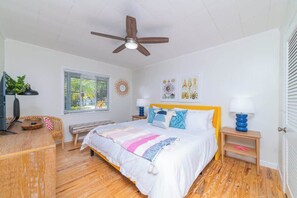 A bedroom with a bed, wooden floor, ceiling fan, bedside tables with lamps, a window with blinds, and a wall decoration.