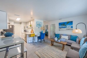 Ein modernes Wohnzimmer mit Sofa, Couchtisch und Essbereich mit Bar. An der Wand hängt ein Gemälde, das eine Strandansicht zeigt.