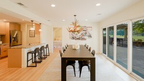 A modern kitchen with a central island, a dining area with a long table, and large sliding glass doors leading to an outdoor patio. - Spacious 5-Bedroom Golf Course Home for Getaways (Merrillville)