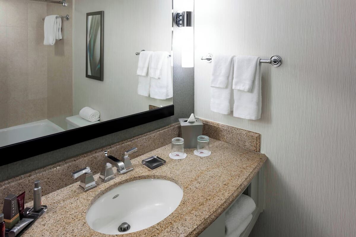 Une salle de bain moderne avec un lavabo doté d’un plan de travail en granit, un grand miroir et des serviettes blanches disposées sur des porte-serviettes.