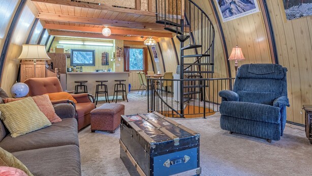 A cozy interior with a curved ceiling, a wooden staircase, a sofa, a recliner, a bar area, and a large window. - A-Frame Cabin nestled in the trees - laundry, pet-friendly, close to village (Silver Star Mountain)