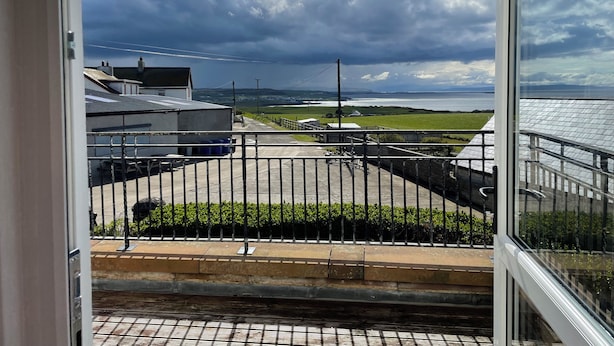 A view from a balcony with a metal railing, overlooking a grassy area and buildings, with a cloudy sky above. - 3 bedroom house (Bushmills)