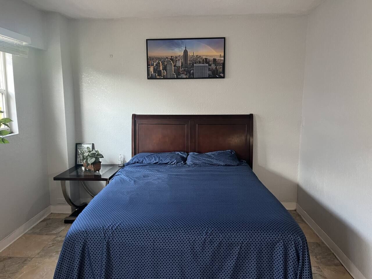 A bedroom with a bed, a nightstand, a framed picture of a cityscape, and a window with a plant.
