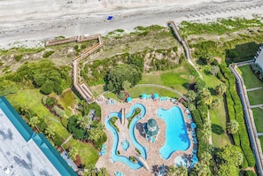 An aerial view of a resort with a large central pool, surrounded by lush greenery and a beach area.