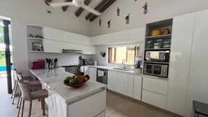 A modern kitchen with white cabinetry, a marble countertop, and a central island with fruit. There are wicker chairs, a coffee machine, and a microwave. - Yeguas V18 by Wynwood House (Anapoima)