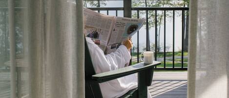 A person reading a newspaper on a wooden deck with a railing, overlooking a body of water and trees.