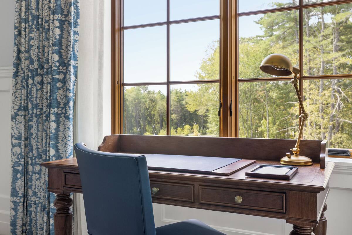 Un bureau en bois, une chaise bleue, une lampe de bureau en laiton et une vue sur les arbres à travers une fenêtre à carreaux.
