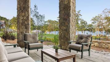 Un patio al aire libre con pilares de piedra, asientos acolchados y vistas a un lago.
