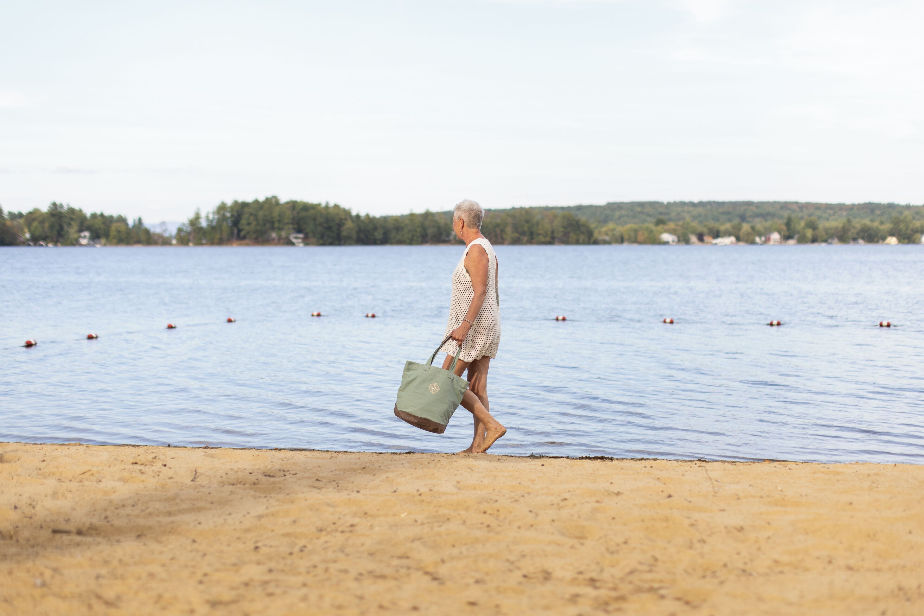 A person walking along a sandy beach with a body of water and trees in the background.