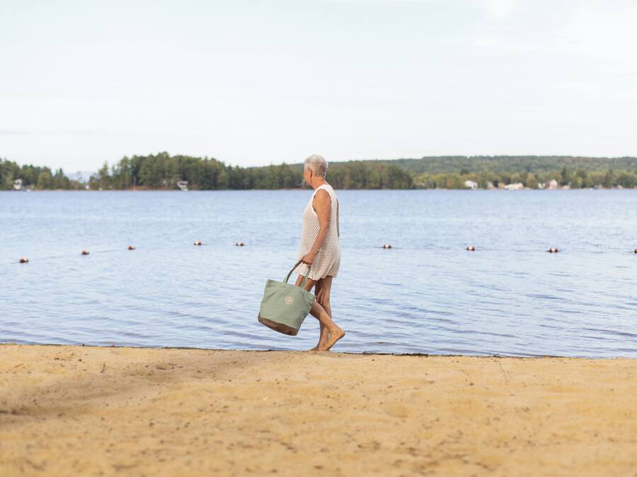 A person walking along a sandy beach with a body of water and trees in the background.