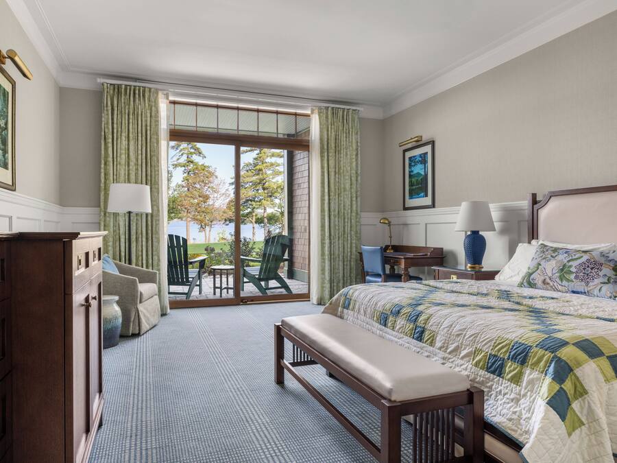 Quarto com cama grande, um banquinho, escrivaninha e uma cadeira. Há vista para o exterior através de uma porta de correr de vidro.