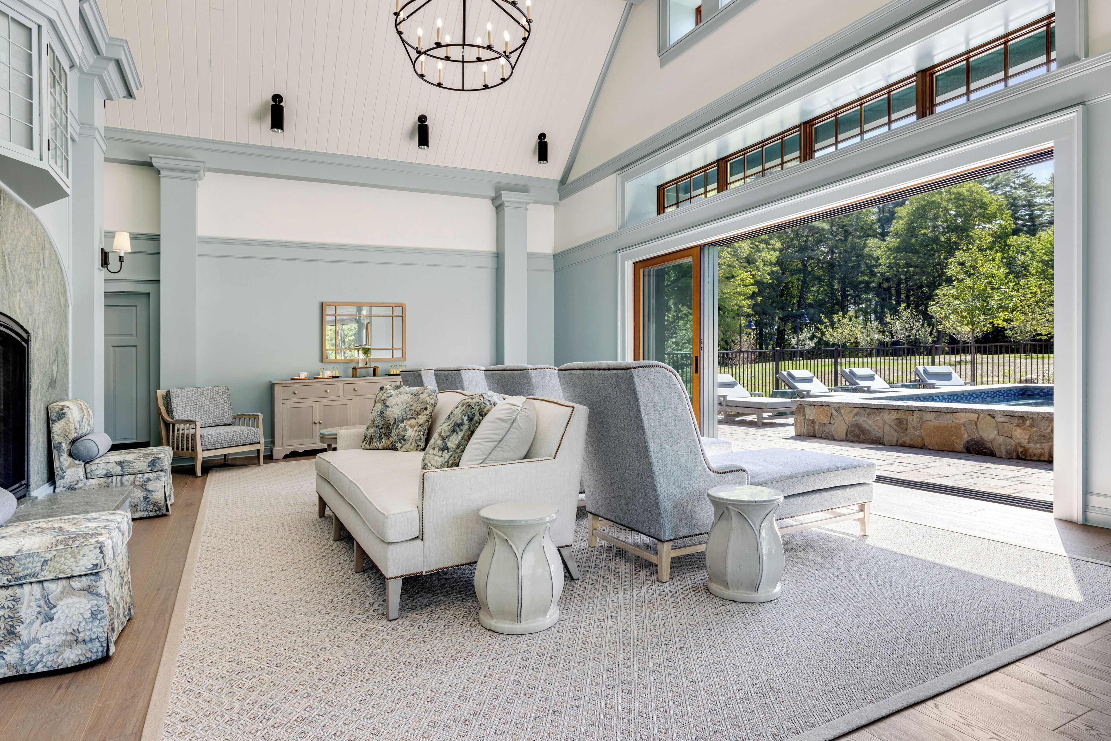 Un salón amplio con chimenea, un sillón gris grande, un sofá blanco y vistas a una piscina y jardín a través de ventanas grandes.