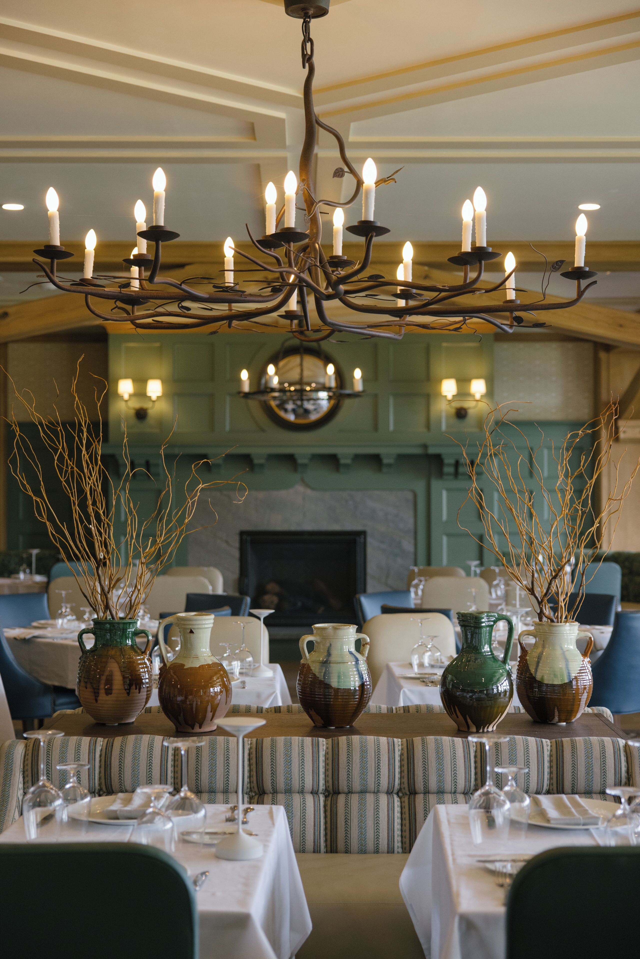 A dining area with tables set for a meal, a chandelier with candles, and vases with branches.