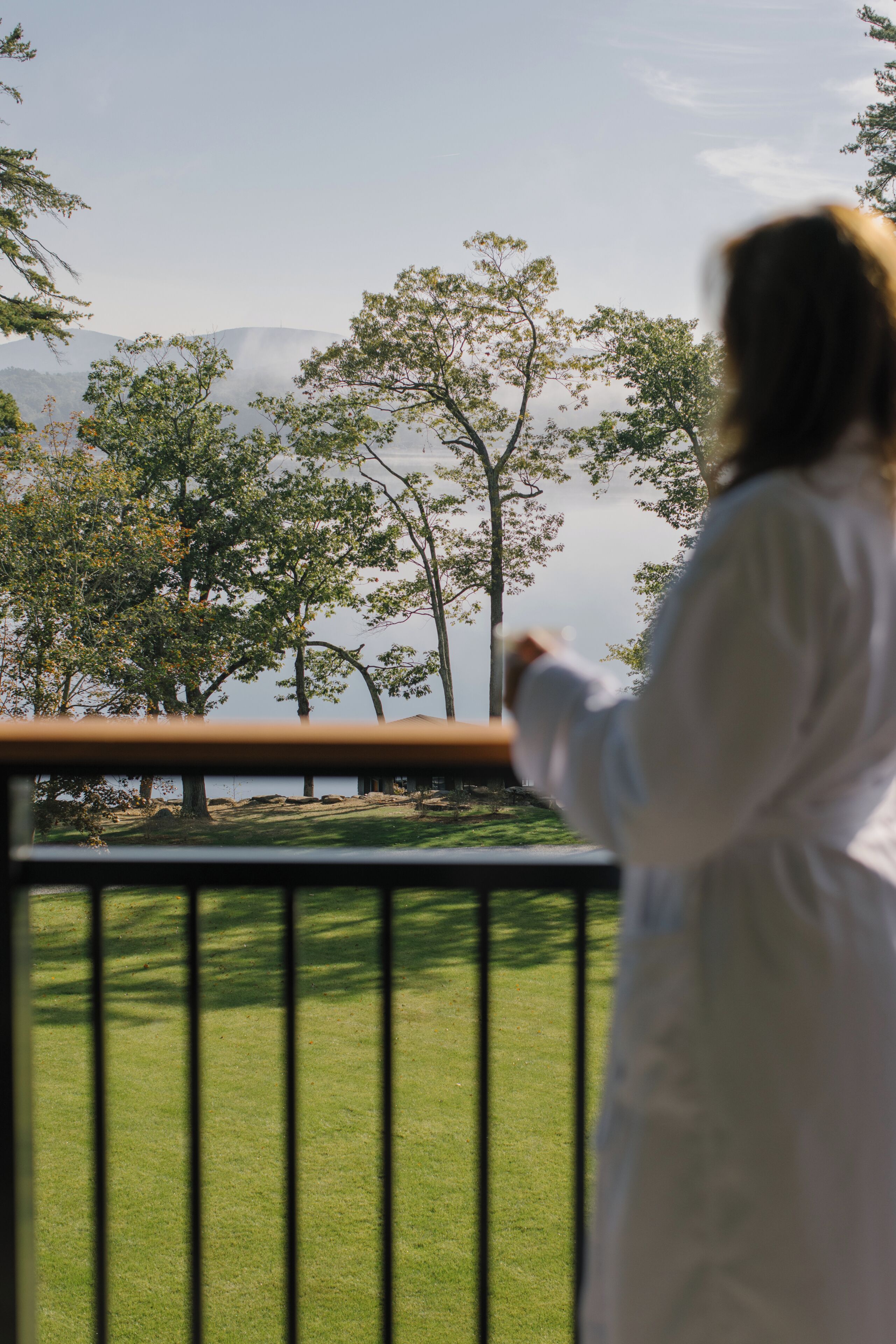 A person in a white coat standing on a balcony with a railing, overlooking a grassy area with trees and a body of water in the distance.