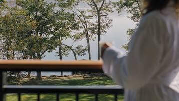 A person in a white coat standing on a balcony with a railing, overlooking a grassy area with trees and a body of water in the distance.