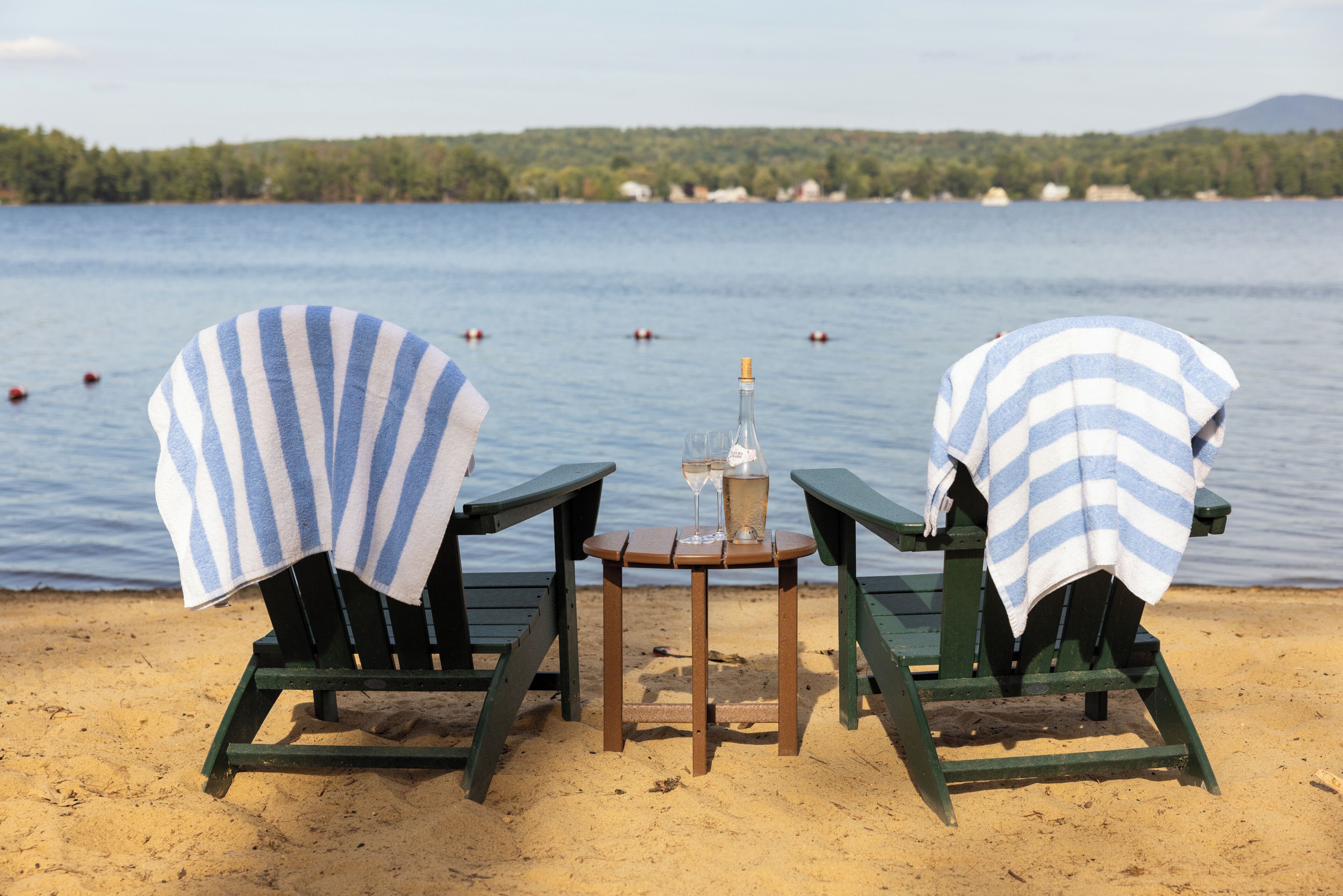 Duas cadeiras de praia com capas listradas, uma mesa pequena com uma garrafa e copos, e vista para um lago com barcos.