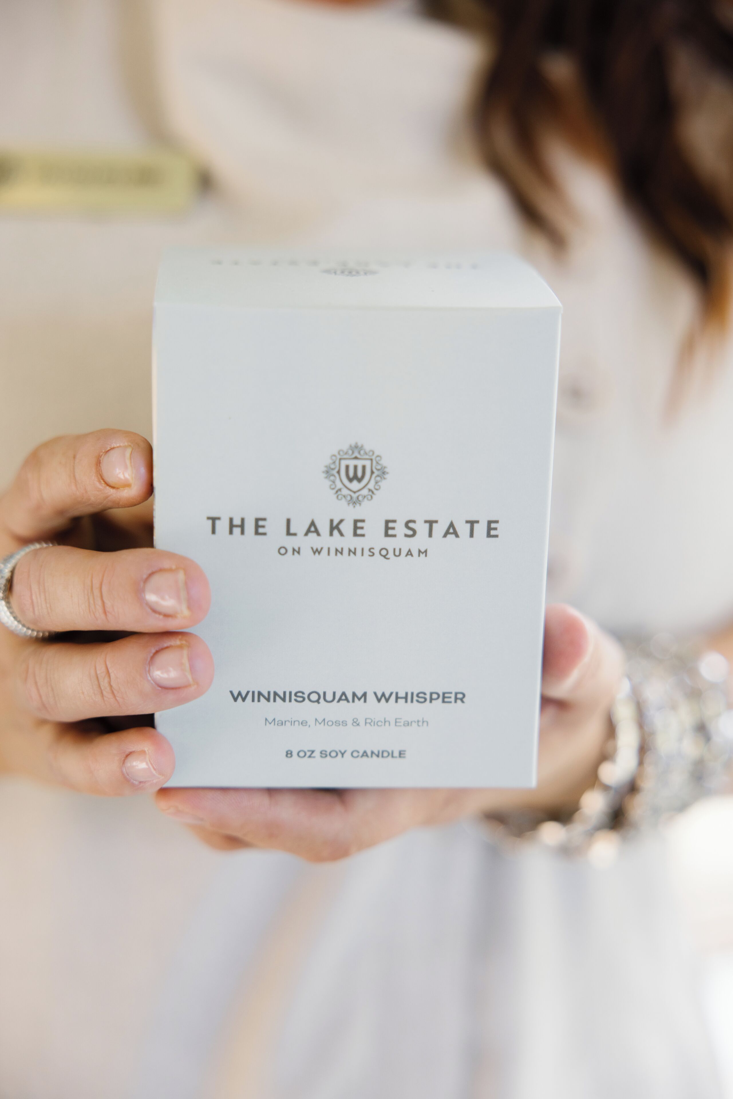 Eine Person hält eine Schachtel, auf der 'The Lake Estate on Winnisquam Whisper 8 oz soy candle' steht.