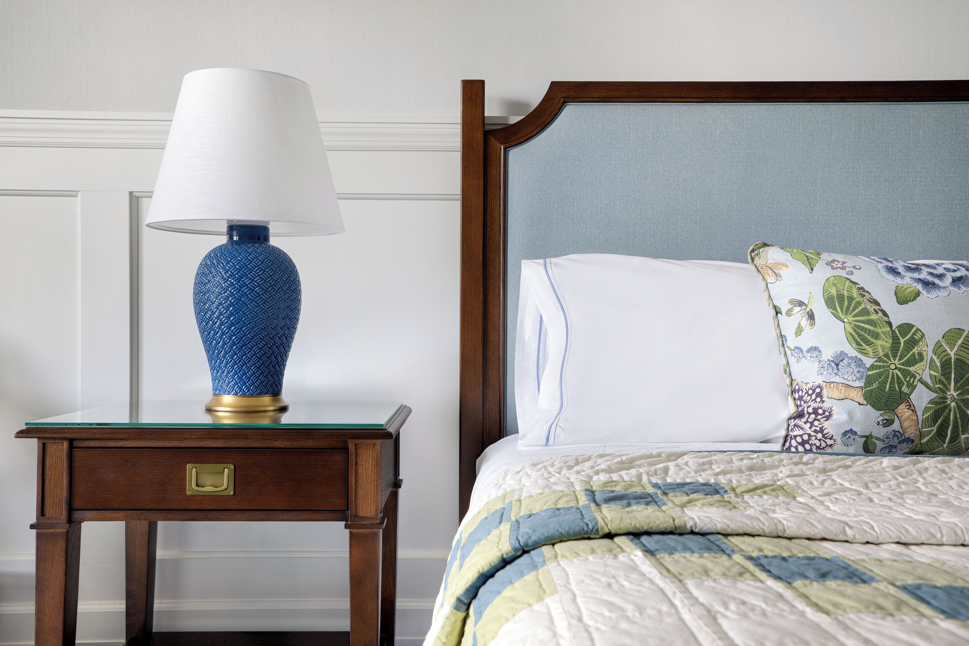 Una cama bien hecha con un cojín floral y una lámpara azul con textura sobre una mesita de noche de madera con un cajón.