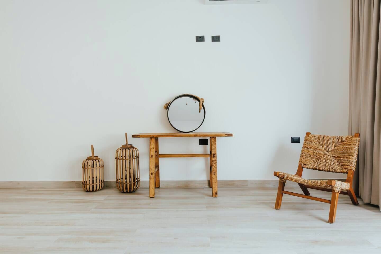 Ett minimalistiskt rum med ett träbord, två lampor av rotting, en flätad stol och en enkel, vit vägg.