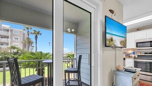 A balcony with chairs, a TV mounted above a kitchen area, and a view of a building and palm trees. - Beachside Villas 613 (Santa Rosa Beach)