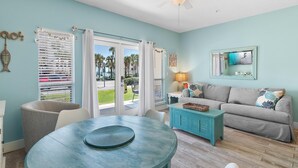 A room with a teal wall, a ceiling fan, a dining table with chairs, a sofa with cushions, and a window with blinds.