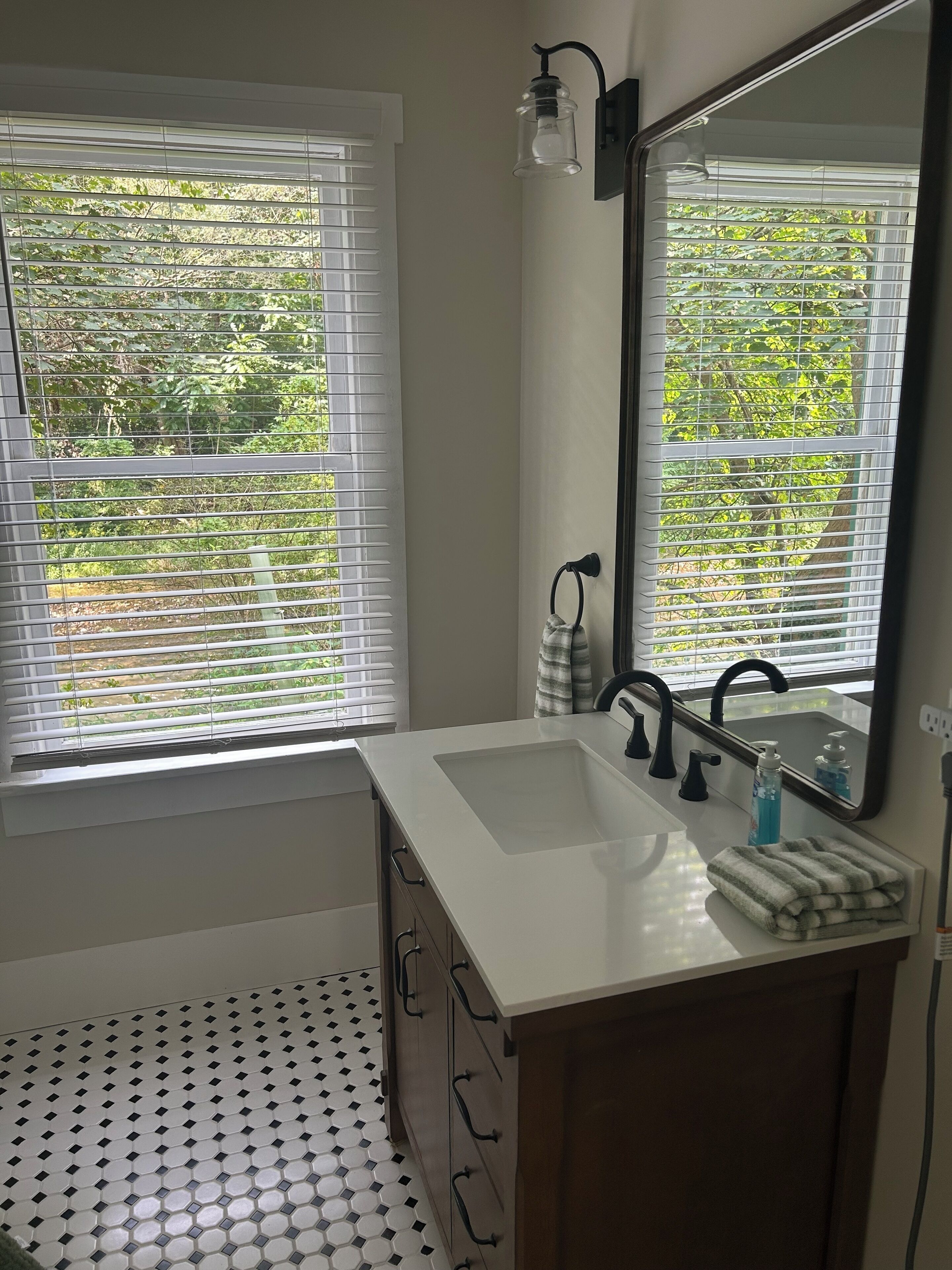 A bathroom with a white sink, black faucet, and a mirror. There are two windows with blinds, and a towel is on the counter.