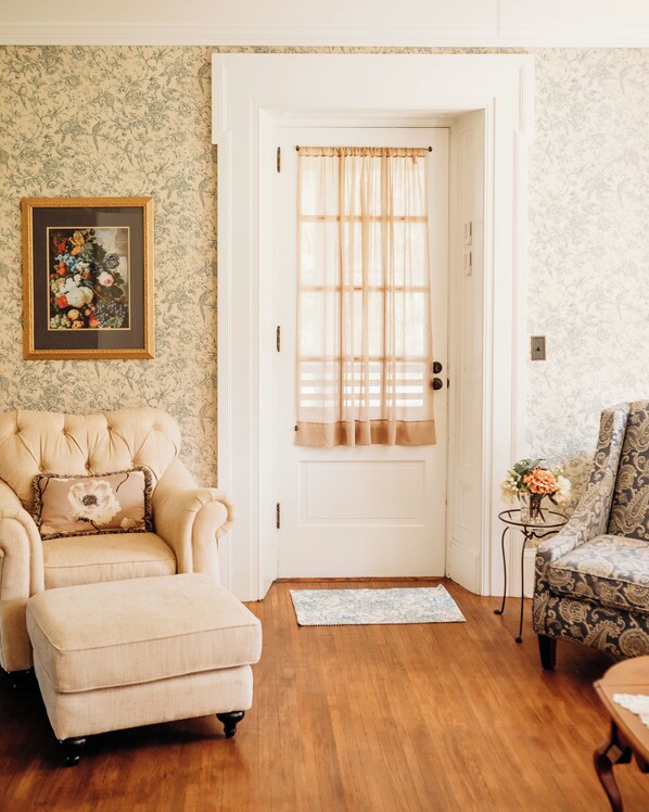 A room with a wooden floor, a white door with a window, a beige armchair with a floral cushion, a patterned armchair, and a small table with a vase of flowers. - Harmony Forge Inn (Milesburg)