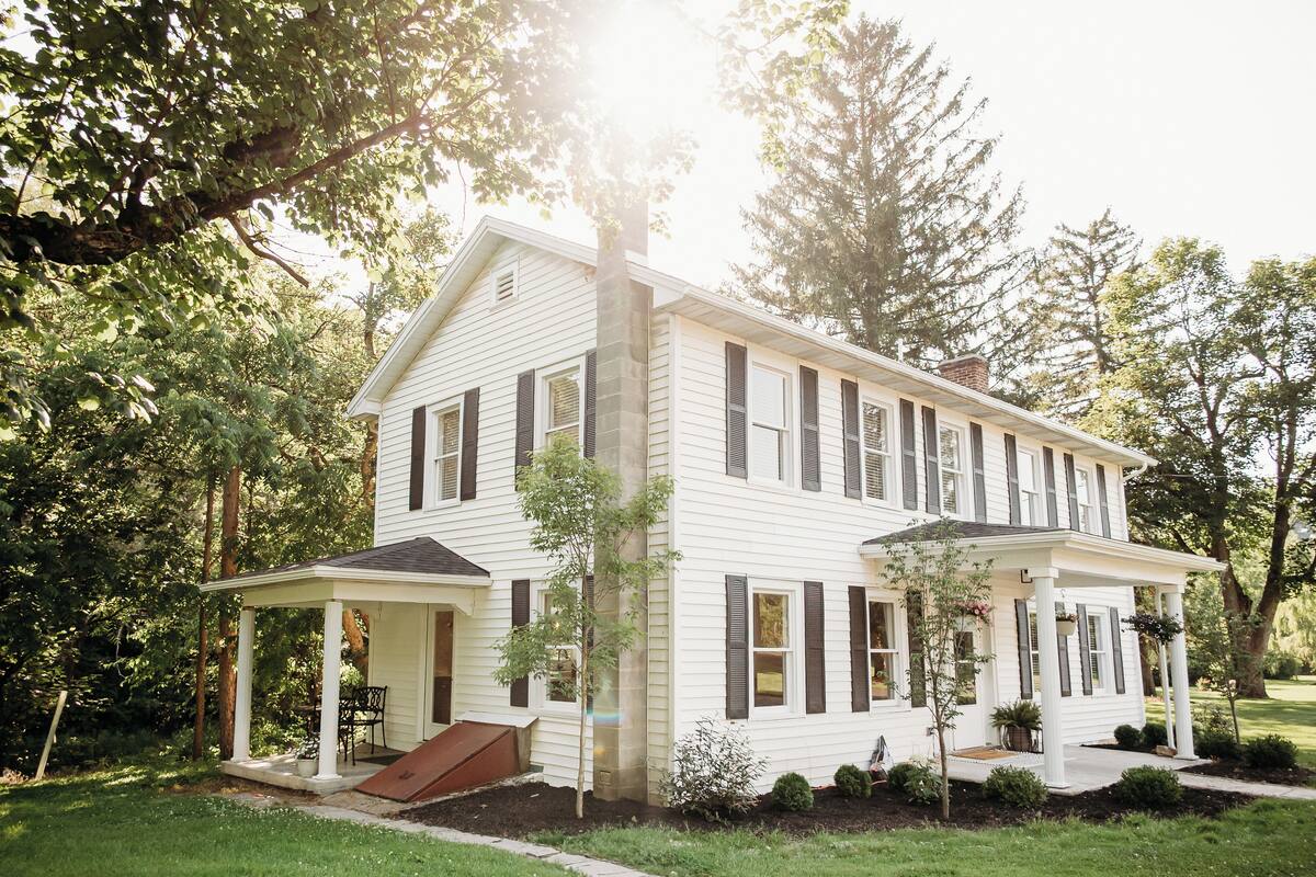 A two-story white house with dark shutters, a covered porch, and a small front yard with trees and shrubs.