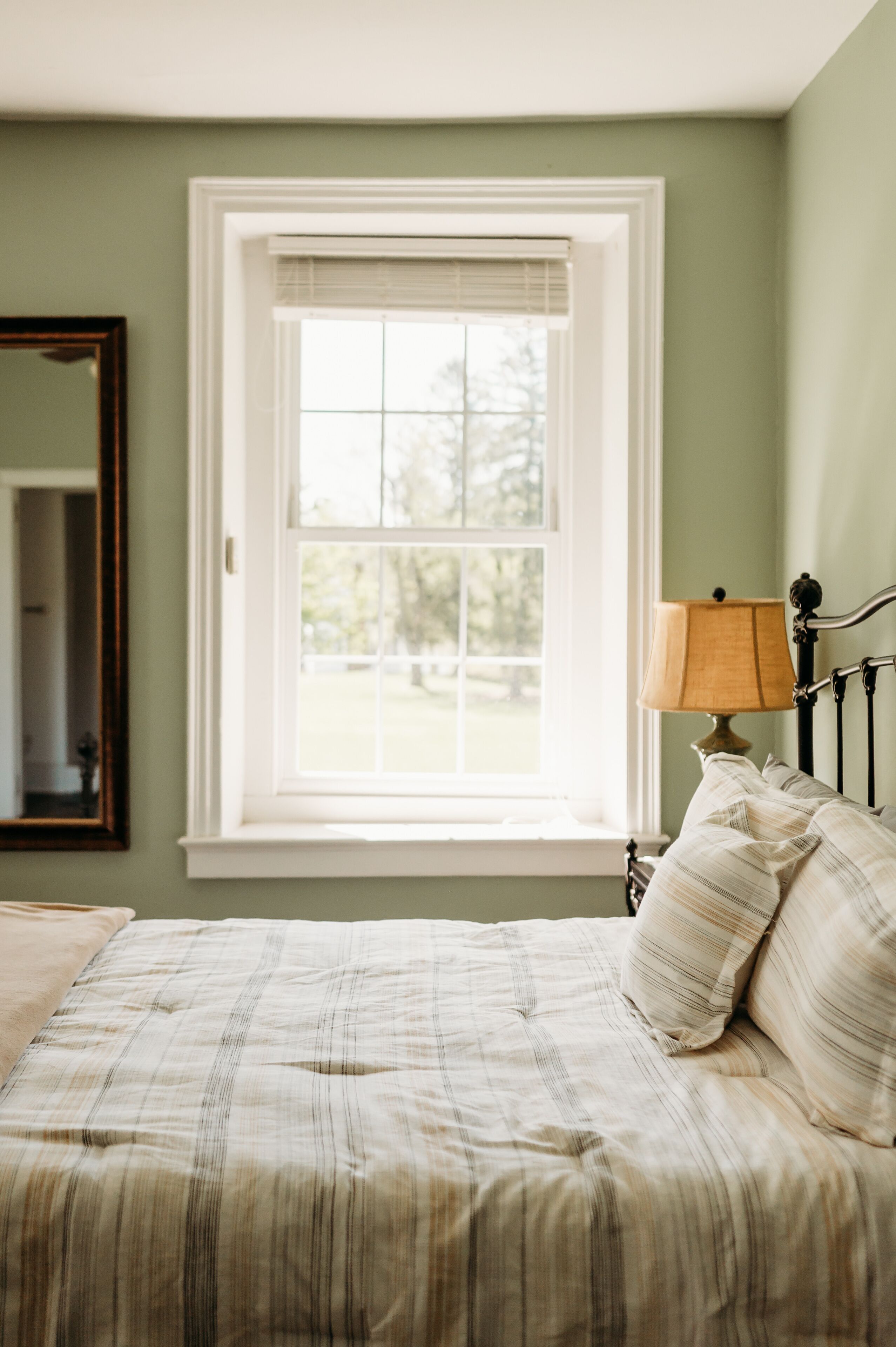 A bedroom with a bed, a window with blinds, a lamp, and a mirror on the wall.