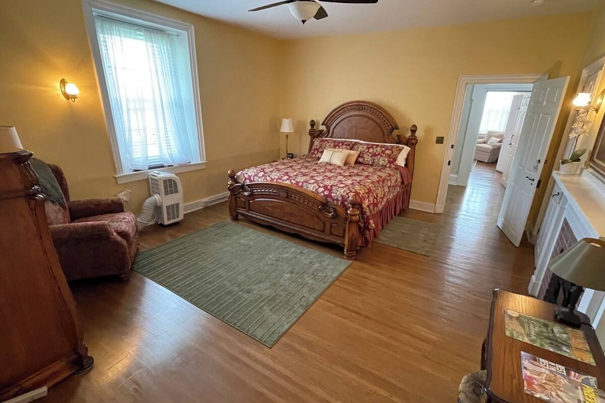 A bedroom with a wooden bed, a chair, a desk, a window with curtains, and a ceiling fan.