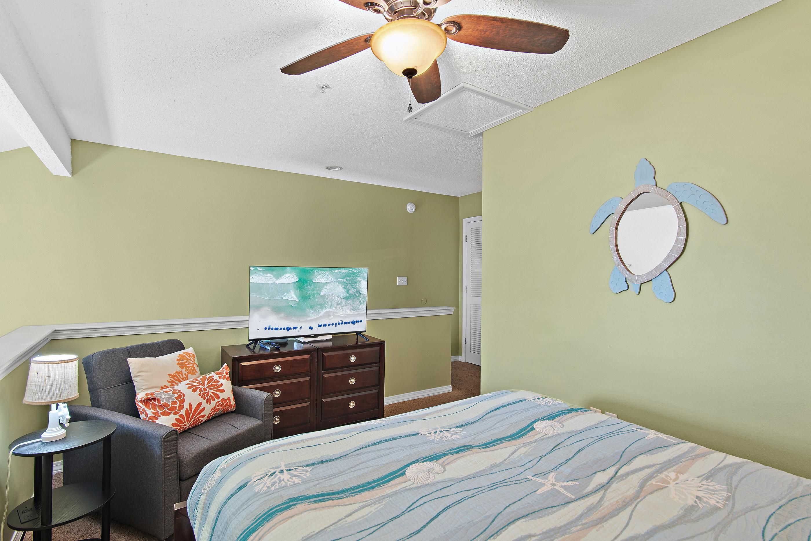 A bedroom with a bed, a television, a dresser, a chair, a lamp, and a ceiling fan.