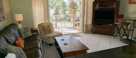 A living room with a sofa, armchair, coffee table, TV stand, and a balcony with a view of palm trees.