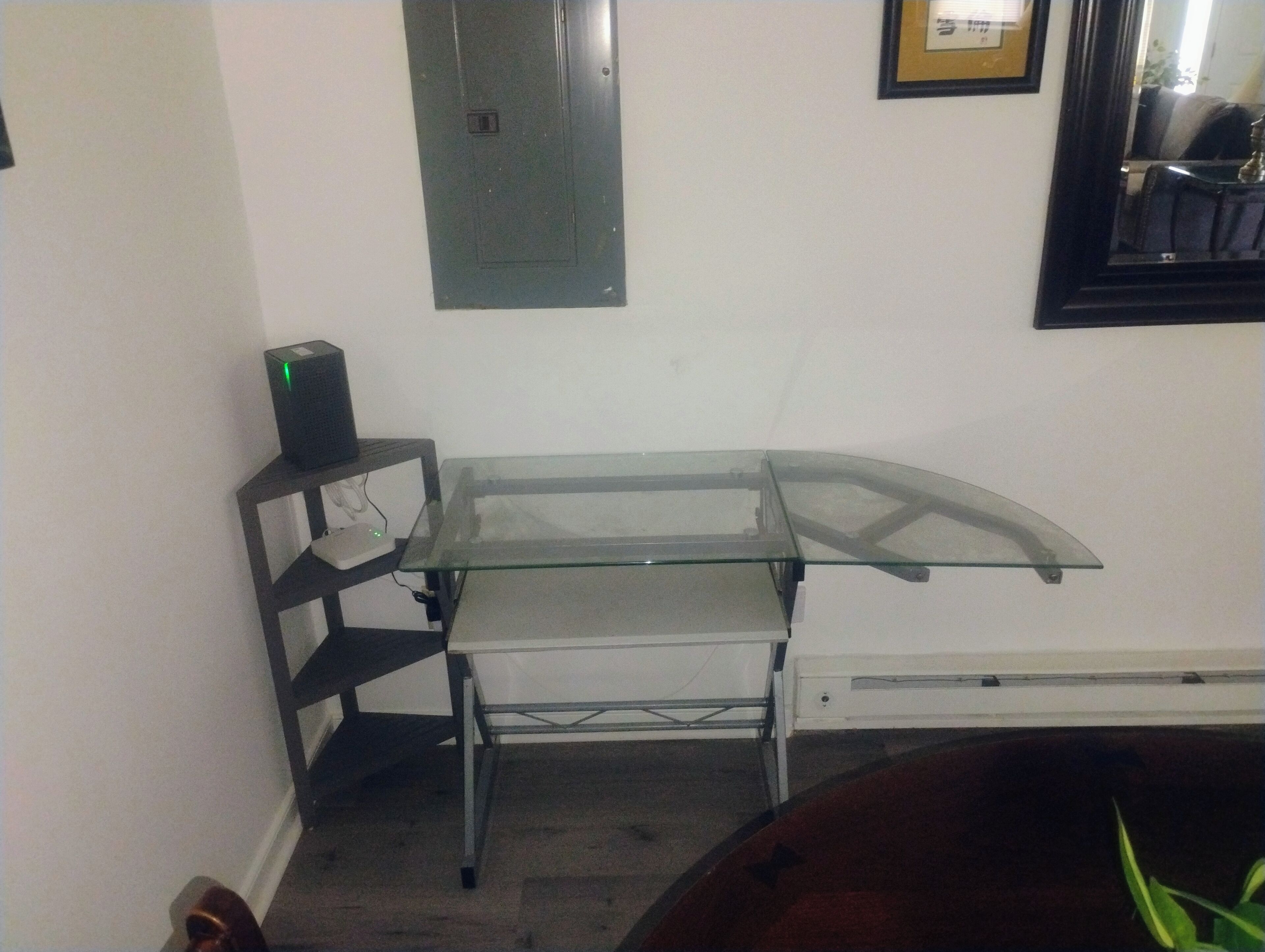 A corner room with a glass-top table, a black speaker on a shelf, and a framed picture on the wall.