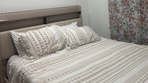 A neatly made bed with striped bedding and decorative pillows, a wall-mounted air conditioner, and a floral-patterned curtain.