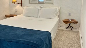 Een slaapkamer met een bed, nachtkastje, lamp, raam met luiken en een klein houten tafeltje.