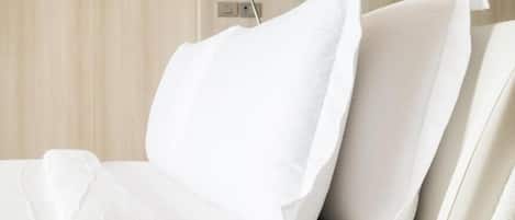 Una cama con sábanas y almohadas blancas, una mesita de noche y un enchufe en la pared.