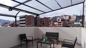 A covered outdoor seating area with a table and chairs, a bottle of soda, and a view of a cityscape with buildings. - Hatun Quilla Apartments (Cusco)