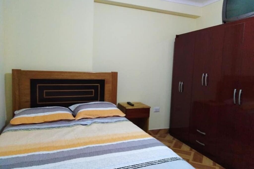 A bedroom with a bed, wooden headboard, striped bedding, a nightstand, a TV, and a wardrobe.
