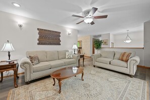 A living room with two sofas, a coffee table, patterned rugs, ceiling fans, wall sconces, and a hanging light fixture.