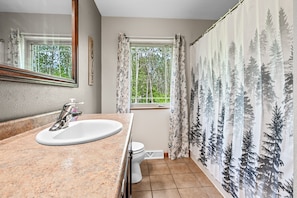 A bathroom with a granite countertop sink, a mirror, a window with curtains, and a shower curtain with a forest pattern.