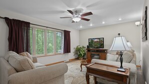 Un salon avec un ventilateur de plafond, un téléviseur, une lampe et une grande fenêtre avec des rideaux.