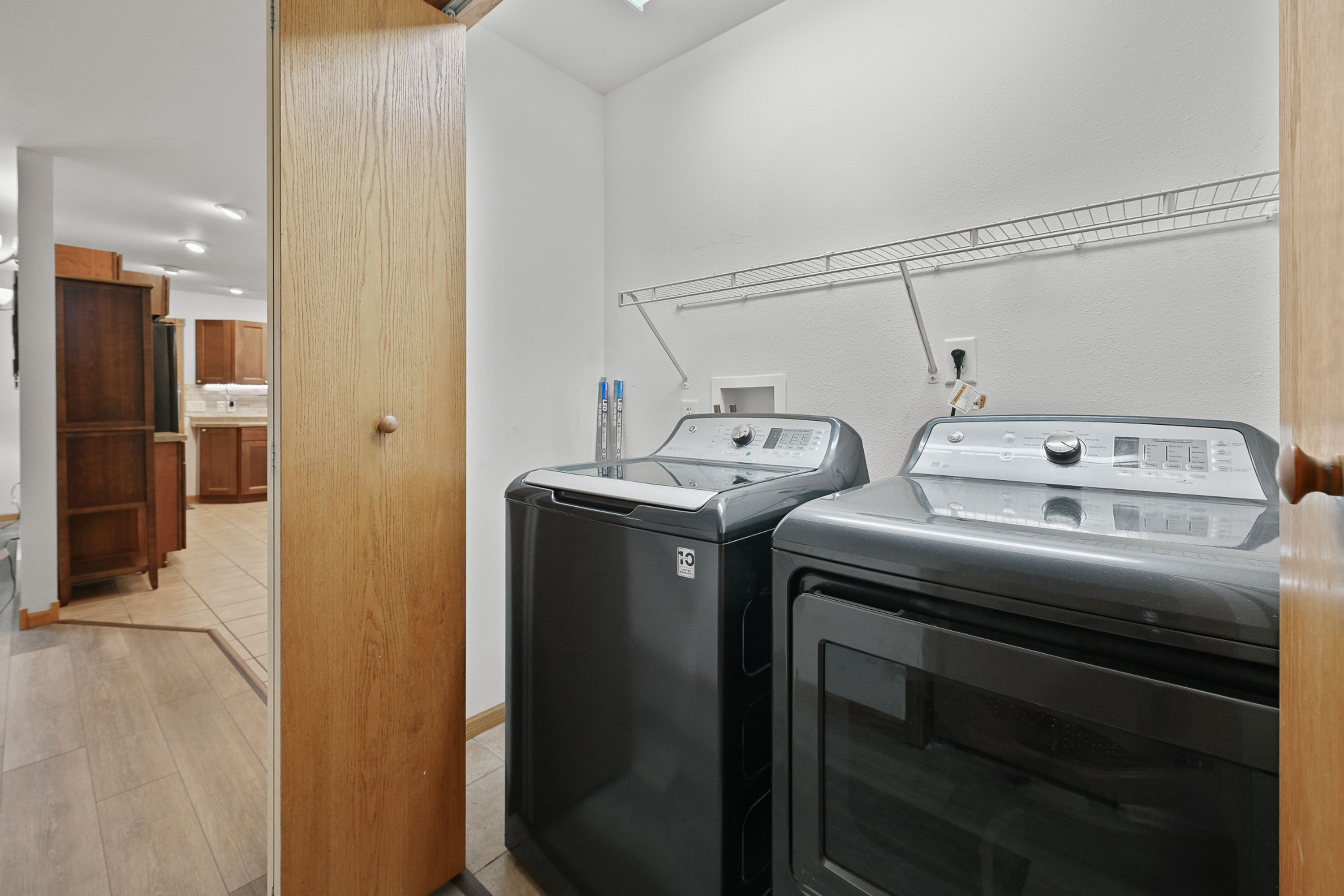 Un espace buanderie équipé de deux machines à laver et d’un sèche-linge, de meubles en bois et d’une étagère en métal placée au-dessus des machines.