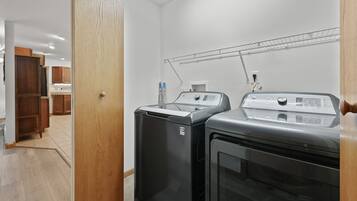Un espace buanderie équipé de deux machines à laver et d’un sèche-linge, de meubles en bois et d’une étagère en métal placée au-dessus des machines.