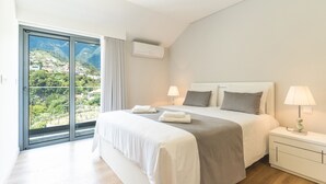 Ein Schlafzimmer mit einem großen Bett, einem Nachttisch, einer Lampe und Blick auf eine gebirgige Landschaft durch eine Schiebetür.