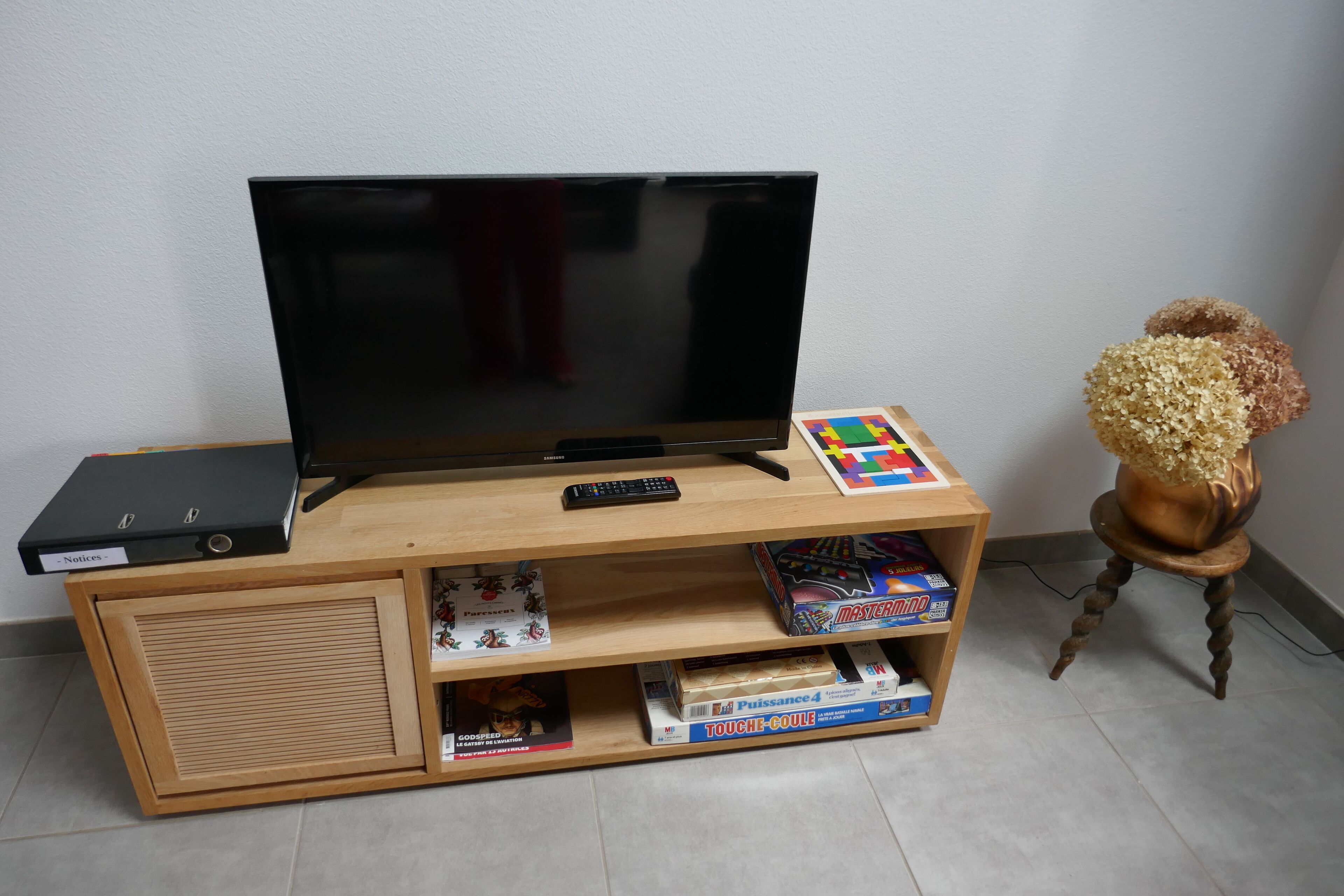 A wooden entertainment center with a flat-screen TV, a remote control, and a small shelf with various items including a game and a box labeled 'TOUCHE-COULE'.