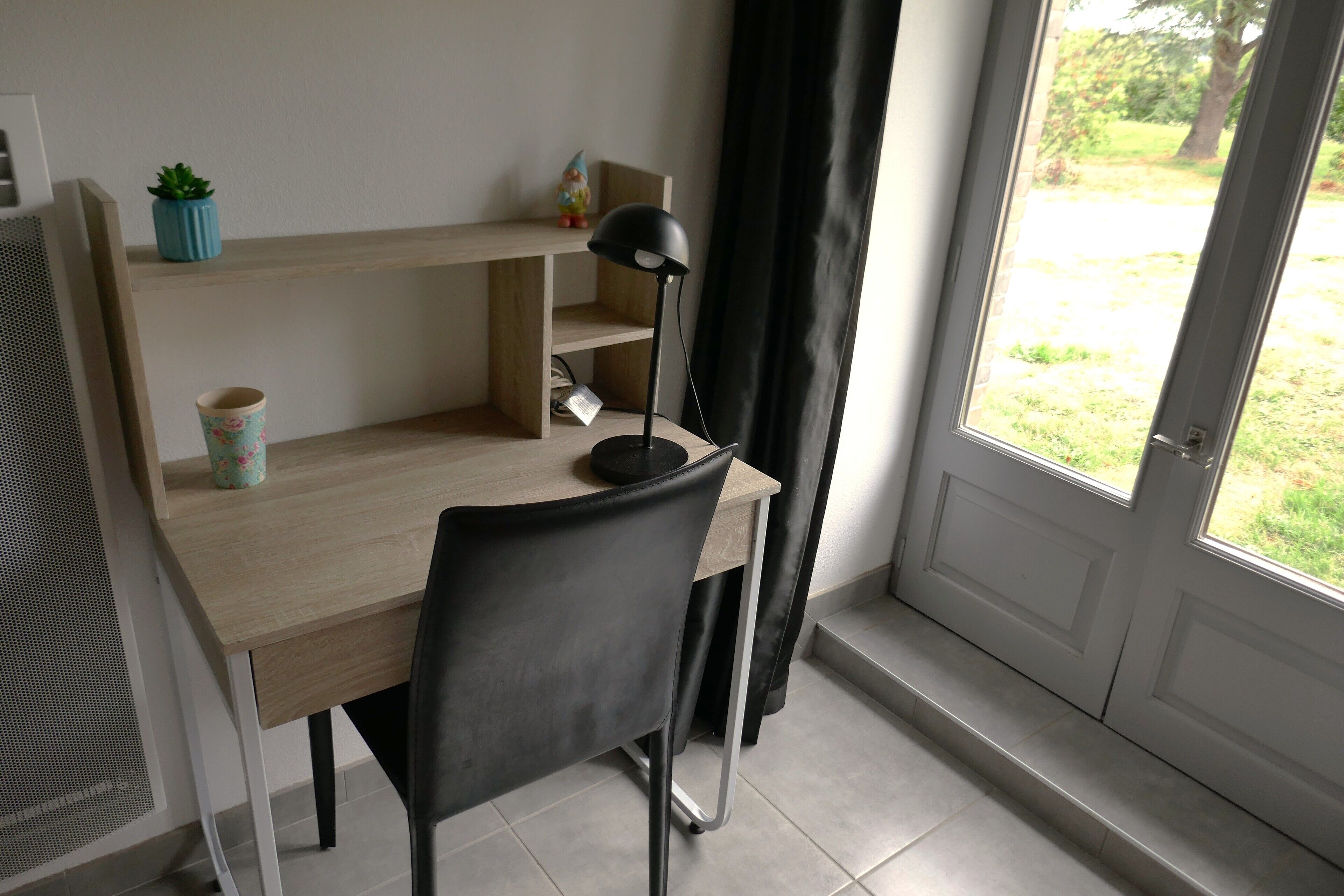 Une pièce avec un bureau en bois, une chaise noire, une petite plante en pot, une lampe et une vue sur l’extérieur, visible à travers une porte.