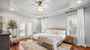 Une chambre à coucher avec un lit en bois, du linge de lit blanc, un ventilateur de plafond et une grande fenêtre avec des stores.